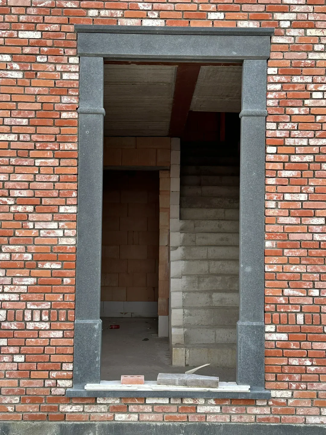 Betonowe schody wewnątrz domu widoczne przez otwór w murze z czerwonej cegły