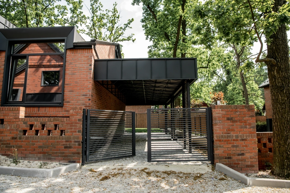 Carport z czarną metalową wiatą i bramą wjazdową na posesji z murem z czerwonej cegły